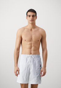Pantaloni de baie albi, cu talie elastică și șnur. Prezintă un logo pe lateral în culorile negru și roșu, confecționați dintr-un material ușor și texturat.
