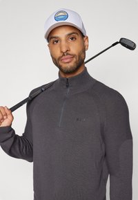 Dunkelgrauer Reißverschluss-Pullover mit strukturierten Schulterakzenten und einem Logo. Weiße Kappe mit einem blauen kreisförmigen Grafik. Golfschläger über der Schulter gehalten.