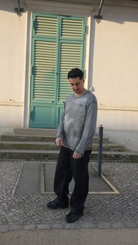 Mladý muž v sivej mikine a čiernych nohaviciach stojí na kockovom chodníku pred zelenými okenicami a kamennými schodmi.