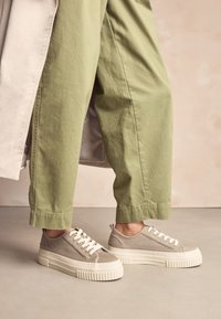 Beige canvas sneakers met een gestructureerd oppervlak, voorzien van witte rubberen zolen en veters, aangevuld met een verhoogd plateau en een lus op de hiel.