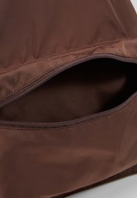 Brun nylonväska med en dragkedjeficka. Tyget har en slät textur och en subtil glans, vilket framhäver dragkedjans metalliska detalj.