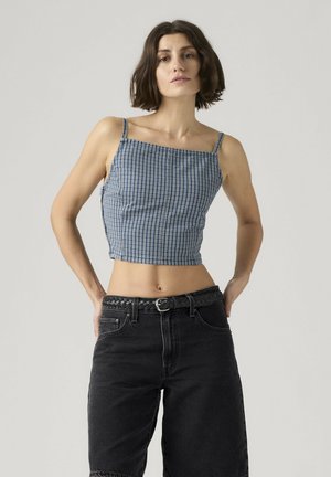Femme aux cheveux courts et foncés portant un crop top à carreaux bleus et un short taille haute noir avec une ceinture tressée, debout devant un fond uni.