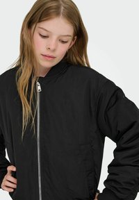 Črna bomber jakna ima zadrgo, rebrast ovratnik in dva stranska žepa. Gladek otip s sproščenim krojem in dolgimi rokavi.