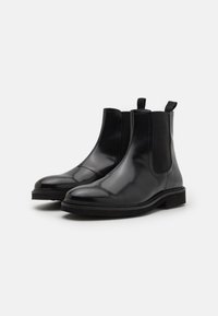 Botoșei Chelsea negri din piele cu un finisaj lucios, cu panouri laterale elastice, tabluță pentru tragere și o talpă groasă neagră. Design durabil și aerodinamic.