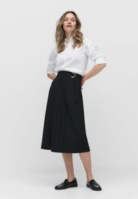 Vrouw staat in een witte lange mouwen-shirt dat in een zwarte geplooide midi-rok is gestoken met een riem en zwarte instappers.