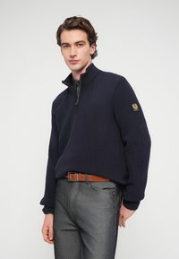 Pull-over marine avec une fermeture éclair d'un quart, texture côtelée et un emblème sur la manche gauche, associé à un jean gris et une ceinture marron.