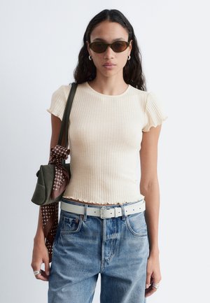 Vrouw met donkere zonnebril, roomkleurige geribbelde korte mouwen top, high-waisted blauwe jeans met witte riem, draagt een groene tas met een patroonachtige sjaal.