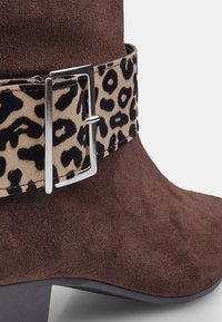 Bottines en suede marron avec une bande en tissu imprimé léopard et des détails en boucle argentée. Design élégant, forme effilée et talon bas.