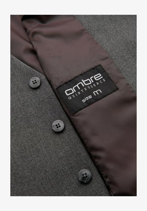 Donkergrijze textuurjas met vier zwarte knopen en een donkerbruine satijnen voering waarop een zwart label staat met 'ombre Quintessence maat M'.