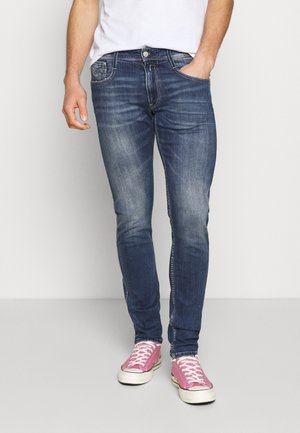 Homme portant un jean bleu délavé, un t-shirt blanc et des baskets basses roses avec des lacets blancs, debout devant un fond clair uni.
