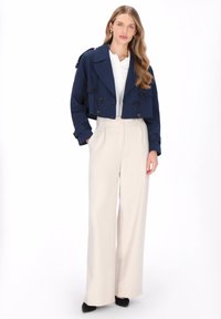 Marinblå kort trenchcoat med dubbelknäppt knäppning, kombinerad med högmidjade creamfärgade vida byxor och svarta högklackade skor. Slät tygstruktur.