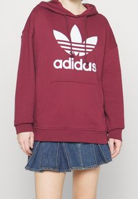 Felpa borgogna con coulisse e un grande logo bianco adidas. Presenta una tasca frontale a marsupio e orlo a coste. Indossata sopra una gonna di jeans.