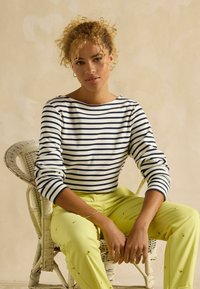 Tumedast ja valgest triibuline laevapitsi särk, millele on kombineeritud erkoranžid püksid, millel on väikesed tikandid. Taustal on punutud tool.