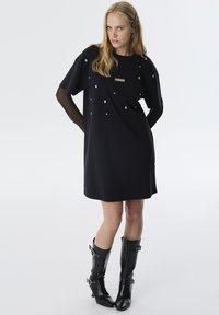 Schwarzes Kleid mit kurzen Ärmeln, das silberne Verzierungen und ein Netzmuster an den Ärmeln aufweist. Kombiniert mit knielangen schwarzen Stiefeln.