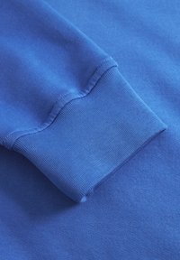 Tissu en coton bleu avec une texture lisse. Le poignet visible présente un design côtelé et des coutures soignées le long des bords.
