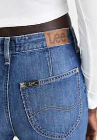 Vue arrière d'une personne portant un jean Lee bleu et un haut blanc à manches longues côtelé, mettant en valeur la poche arrière et les écussons du logo du jean.