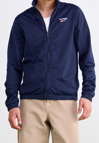 Laivastonsininen vetoketjullinen urheilu-takki, joka on valmistettu sileästä, kevyestä kankaasta. Takissa on Reebokin logo rinnassa, joustavat hihansuut ja helma.