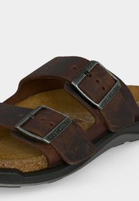 Καφέ δερμάτινο σανδάλι με δύο ρυθμιζόμενες μεταλλικές αγκράφες χαραγμένες με την επιγραφή "BIRKENSTOCK" και φελλό πέλμα σε μαύρη σόλα.