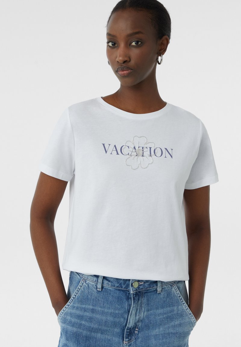 T-shirt en coton blanc à manches courtes, avec un motif floral "VACANCES" en broderie bleue et argentée sur le devant.