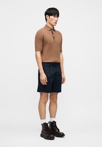 Καφέ ρι γωτό polo με κοντά μανίκια, σκούρο μπλε σορτς και καφέ δερμάτινα μπότες με κορδόνια και λευκές κάλτσες. Απλός σχεδιασμός και εφαρμοστό περίγραμμα.