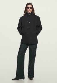 Cappotto di lana nero con collo alto, chiusura a bottoni e tasche laterali. Abbinato a pantaloni di denim verde scuro e scarpe nere a punta.