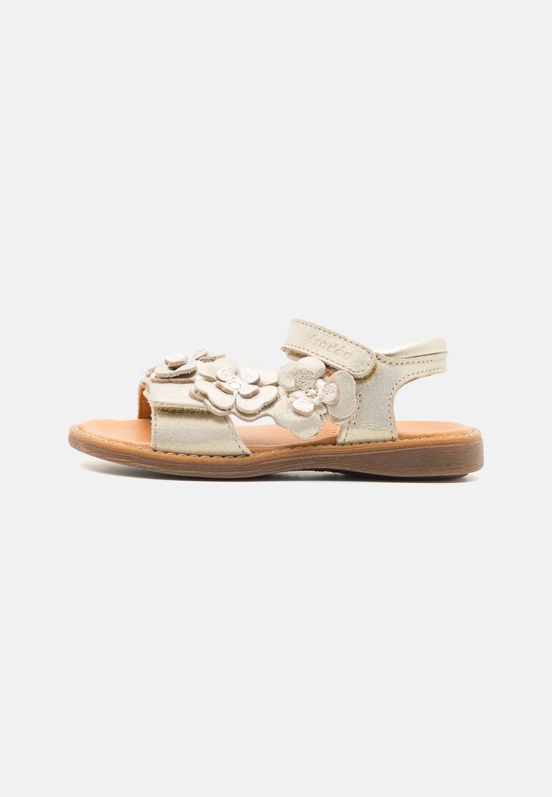 Sandal bej deschis cu flori, realizat din suede, cu o talpă plată din cauciuc. Prezintă vârful deschis, curea ajustabilă și accesorii decorative sub formă de flori.