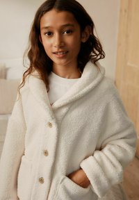 Mädchen mit langen Haaren, das eine weiße, flauschige Jacke mit Knöpfen und Taschen trägt, steht drinnen mit einer Hand in der Tasche.