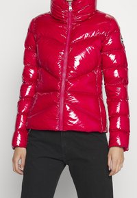 Skinande, isolerad puffjacka i starkt rosa med hög krage och blixtlås framtill. Har ett chevron-quiltmönster och elastiska muddars.