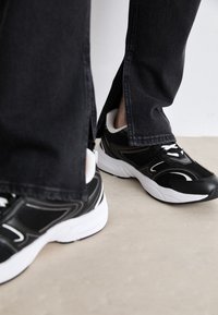 Black athletic shoes with white accents and mesh panels, paired with dark denim jeans featuring side slits. Smooth rubber soles.