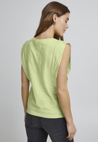 Top senza maniche di un verde chiaro con una vestibilità rilassata, texture morbida e bordi grezzi. Presenta un scollo rotondo e un design posteriore leggermente largo.