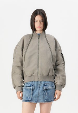 Giovane donna con i capelli scuri e lisci, che indossa una giacca bomber grigia oversize e una gonna di jeans blu con grandi tasche frontali.