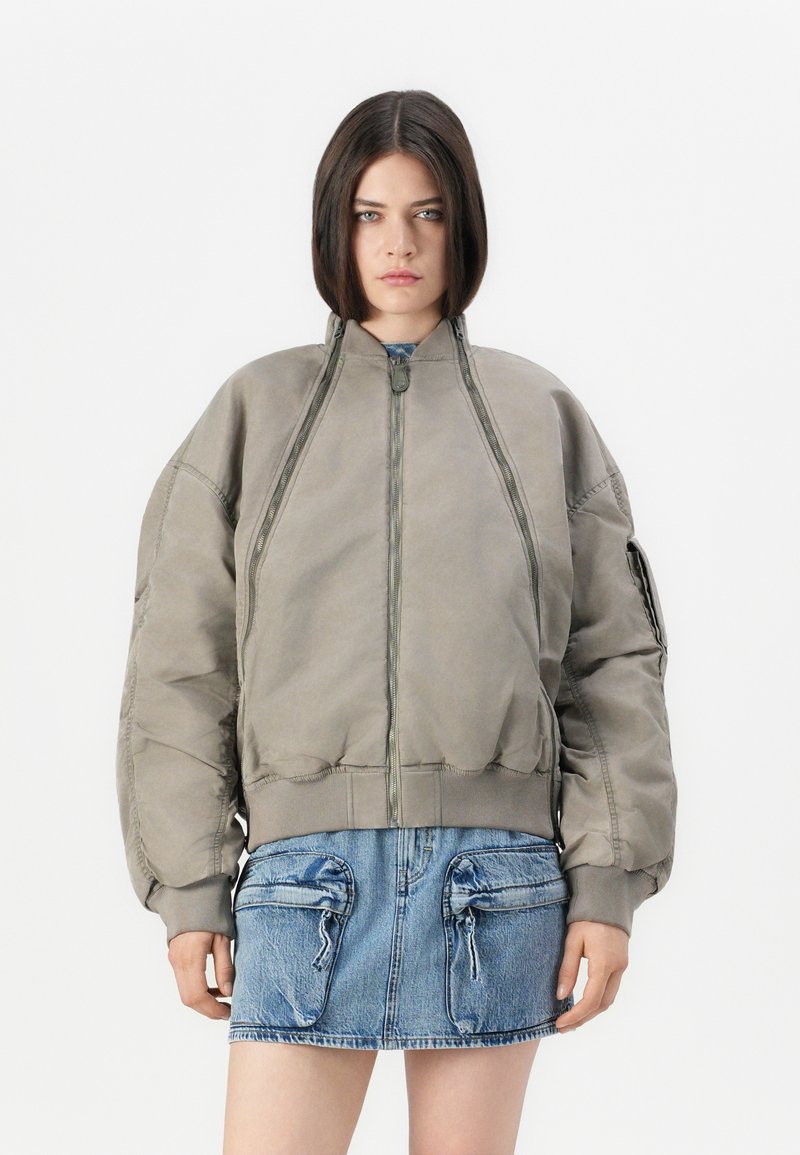 Giovane donna con i capelli scuri e lisci, che indossa una giacca bomber grigia oversize e una gonna di jeans blu con grandi tasche frontali.
