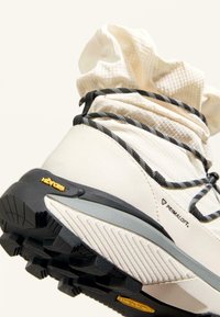 Krémové boty s texturovaným svrškem, černobílými tkaničkami a Vibram podešví. Obsahují branding Primaloft a polstrovaný límec kolem kotníku.