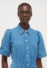 Blå denimklänning med korta puffärmar, krage och detaljer i guldknappar. Har vertikal sömnad och en figursydd midjedesign.