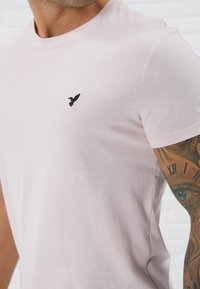 Camiseta de algodón de color rosa claro con mangas cortas, presenta un logotipo de pájaro bordado en negro en el pecho, textura suave y un cuello redondo clásico.