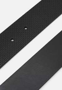 Cintura nera con un motivo testurizzato a griglia su un lato e una finitura liscia dall'altro. Presenta due fori per la regolazione.