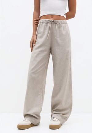Kobieta w beżowych szerokich spodniach sztruksowych z wiązanym paskiem w talii i białych trampkach, w połączeniu z białym, krótkim topem na ramiączkach.