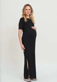 Schwarzes, bodenlanges Umstandskleid mit kurzen Ärmeln und seitlichem Schlitz. Glattes Material mit taillierter Passform, getragen mit gemusterten High Heels.