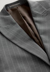 Chaqueta de traje gris con finas rayas verticales, que cuenta con un forro de satén marrón. Los detalles notables incluyen solapas afiladas y un bolsillo.