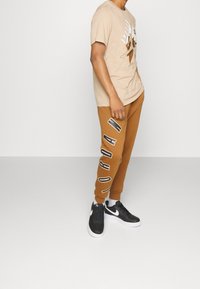 Man som bär en beige Jordan-t-shirt, bruna Jordan-mjukisbyxor med stort logo på benet och svarta Nike-sneakers med vita sulor.