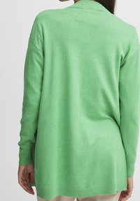 Kobieta ubrana w luźny, jasnozielony sweter z długim rękawem, pokazana od tyłu, w beżowych spodniach i z długimi ciemnymi włosami.