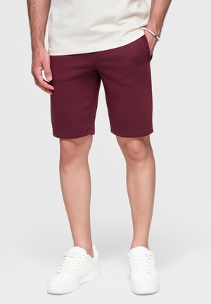 Homme portant un short bordeaux mi-cuisses, des baskets blanches et une chemise blanche, une main dans la poche et un bracelet bleu à perles.