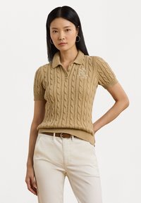 Kabelgebreide, kortegalpolo in beige met een kraag en geborduurd logo. Gecombineerd met crèmekleurige broek en een bruine riem.