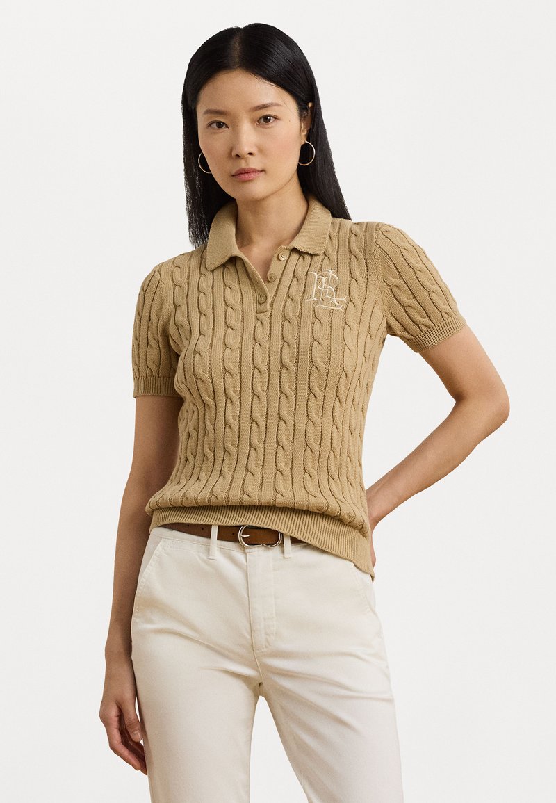 Kabelgebreide, kortegalpolo in beige met een kraag en geborduurd logo. Gecombineerd met crèmekleurige broek en een bruine riem.