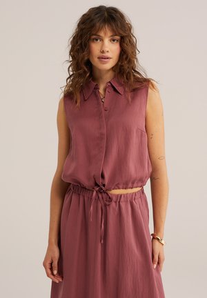 Femme aux cheveux bouclés portant un haut mauve sans manches boutonné, noué à la taille, et une jupe taille haute assortie, debout sur fond uni.