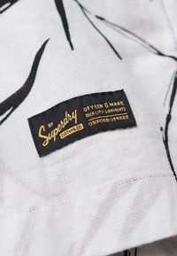 Tela blanca con patrones abstractos en negro. Presenta una etiqueta negra con texto dorado que dice "Superdry" y "Styled & Made Quality Garments Oxford Street".