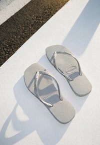 Paire de tongs Havaianas gris clair avec semelles texturées posée sur une surface blanche, la lumière du soleil projette des ombres en angle.