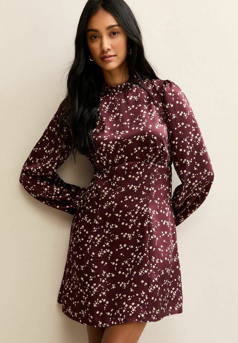 Vrouw draagt een lange mouw, bordeauxrode jurk met een wit bloemenpatroon, staand tegen een effen beige muur met haar handen achter haar rug.