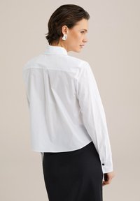 Camisa larga de algodón blanca de manga larga con dobladillo curvado, pliegue trasero y puños abotonados, combinada con una falda negra y pendientes simples.