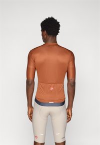Maillot de cyclisme marron, ajusté, avec des manches courtes, tissu texturé et une poche arrière. Associé à un short beigeon rembourré avec une ceinture noire.